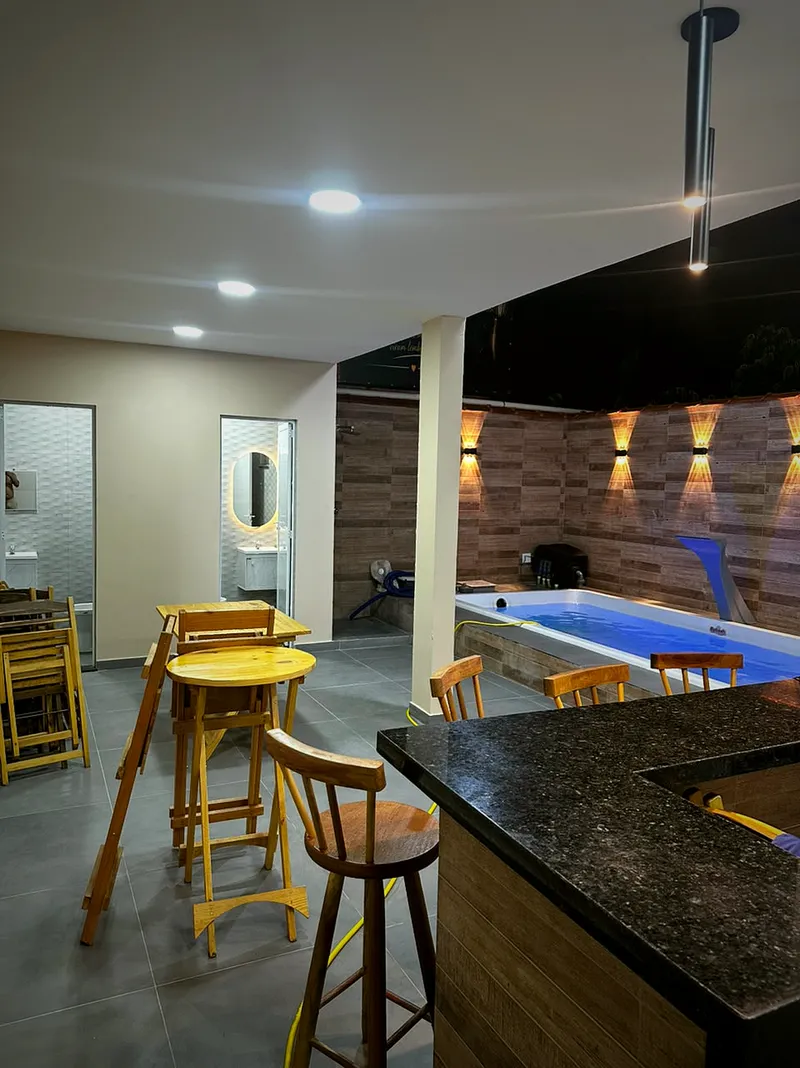 Área coberta com bancada de granito, banquetas de madeira, mesas e a piscina ao fundo